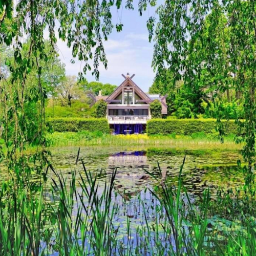 Photo of LongHouse Reserve, a building with an A-frame roof tucked behind a series of trees and hedges, with its facade overlooking a mossy and lilypad-laden pond.